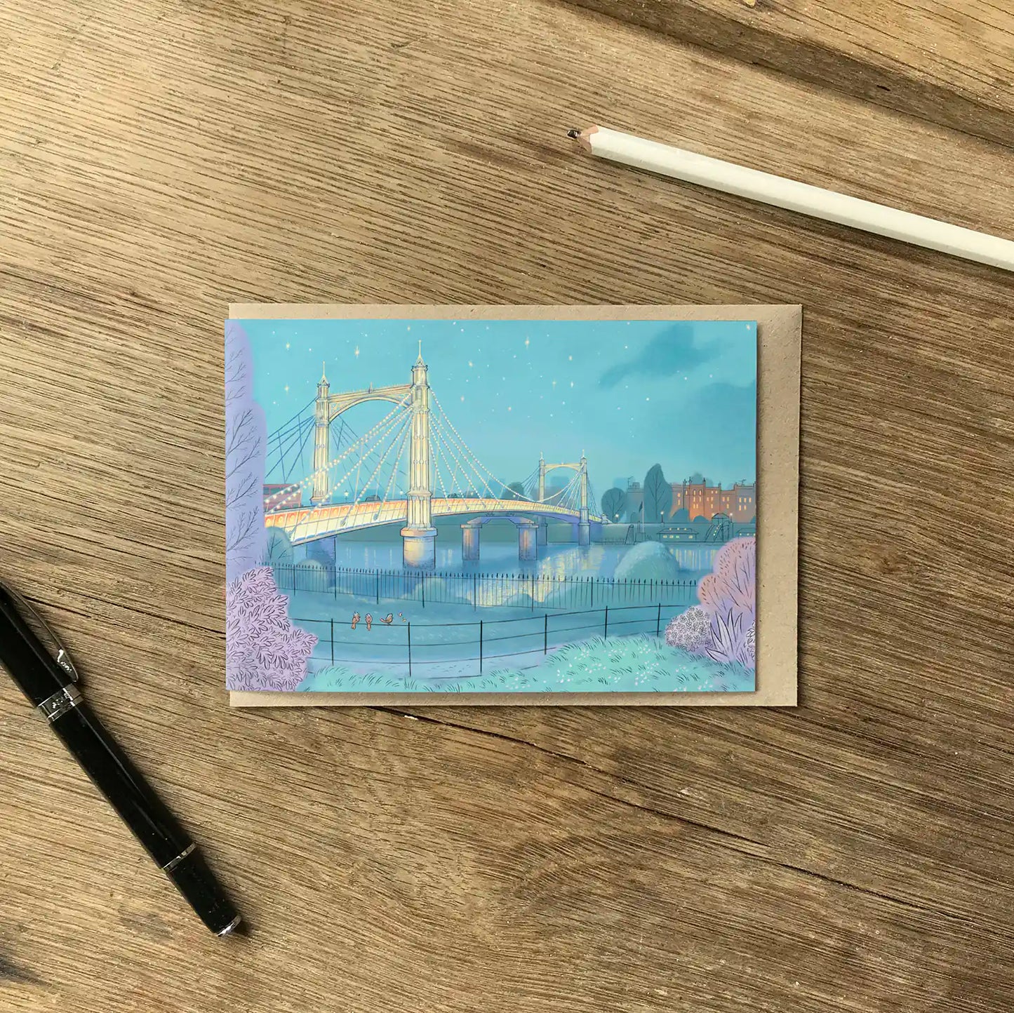 Greetings card of Albert Bridge on the Thames London painted by Mike Green Illustration on a wooden table