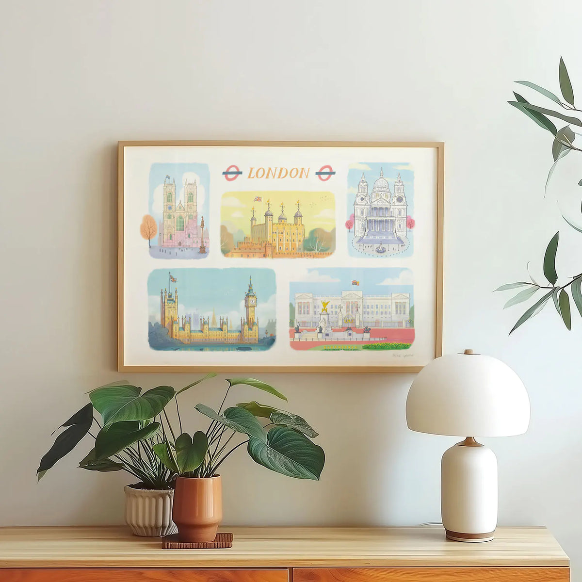 A framed art print of London's landmarks including, Westminster Abbey, The Tower of London, St Paul's Cathedral, Palace of Westminster and Buckingham Palace on a wall with a plant and lamp