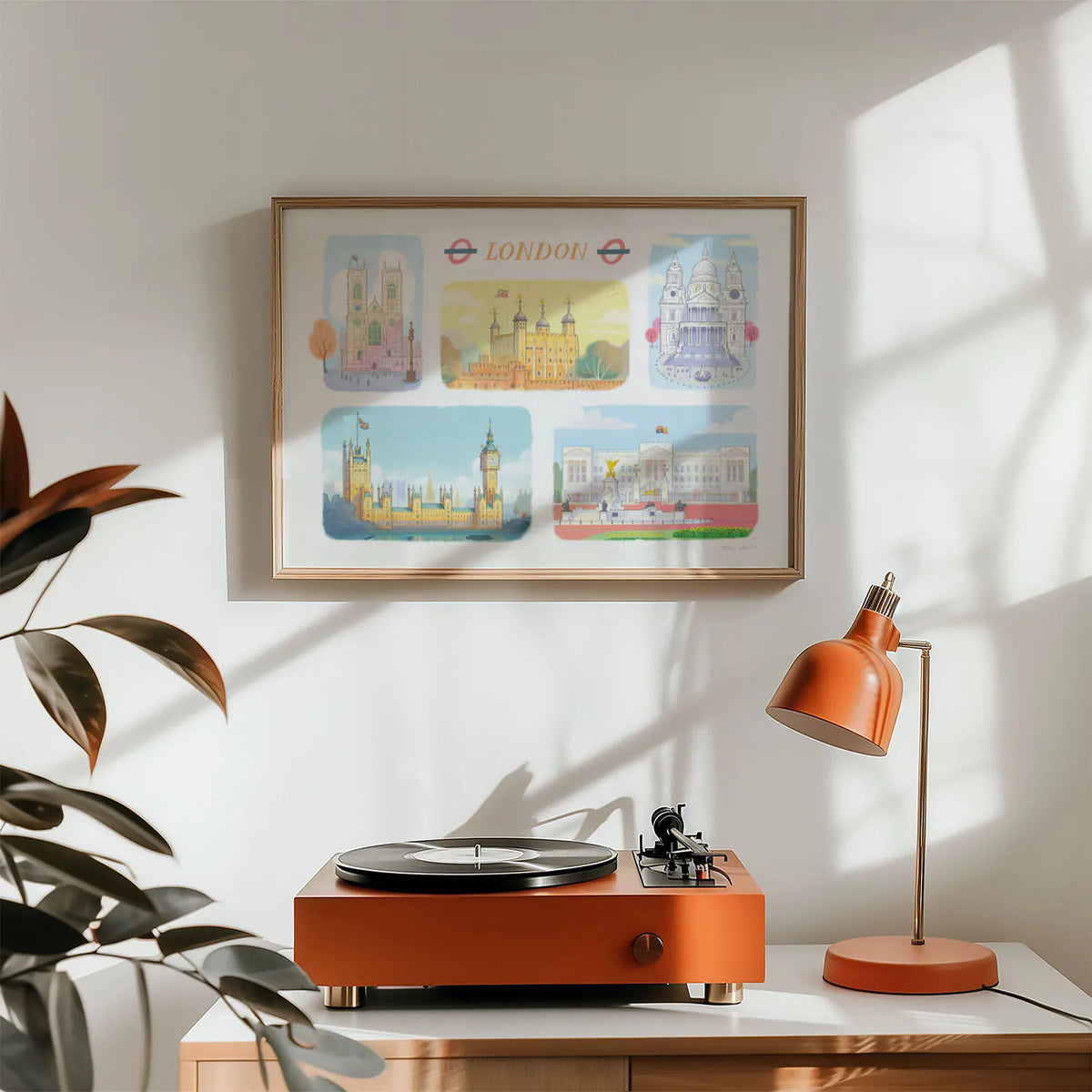 A framed art print of London's landmarks including, Westminster Abbey, The Tower of London, St Paul's Cathedral, Palace of Westminster and Buckingham Palace on a wall with a record player and lamp
