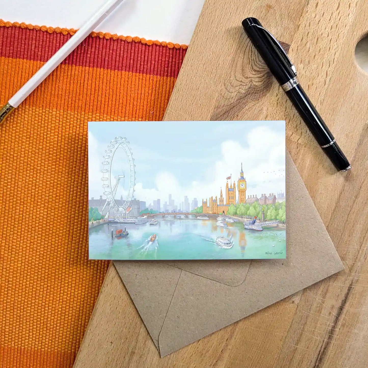 Greetings card on desk with pens of an illustration of the view south from London's Golden Jubilee Bridge with Houses of Parliament and the London eye by Mike Green Illustration