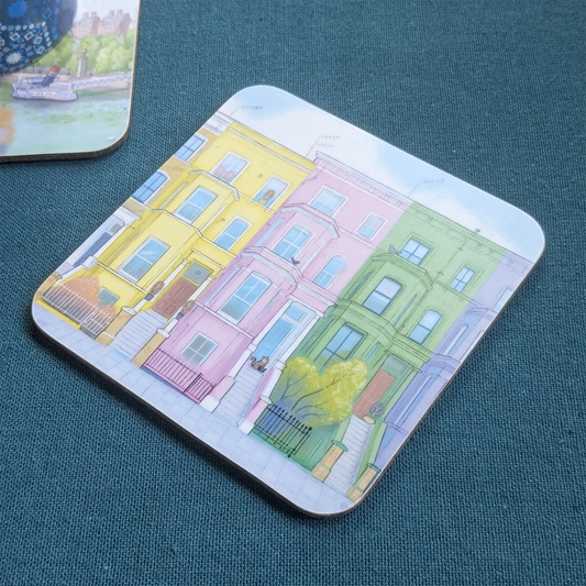 A square coaster featuring an illustration of colorful houses in London's Notting Hill with a cat, placed on a textured dark teal surface next to a knife.