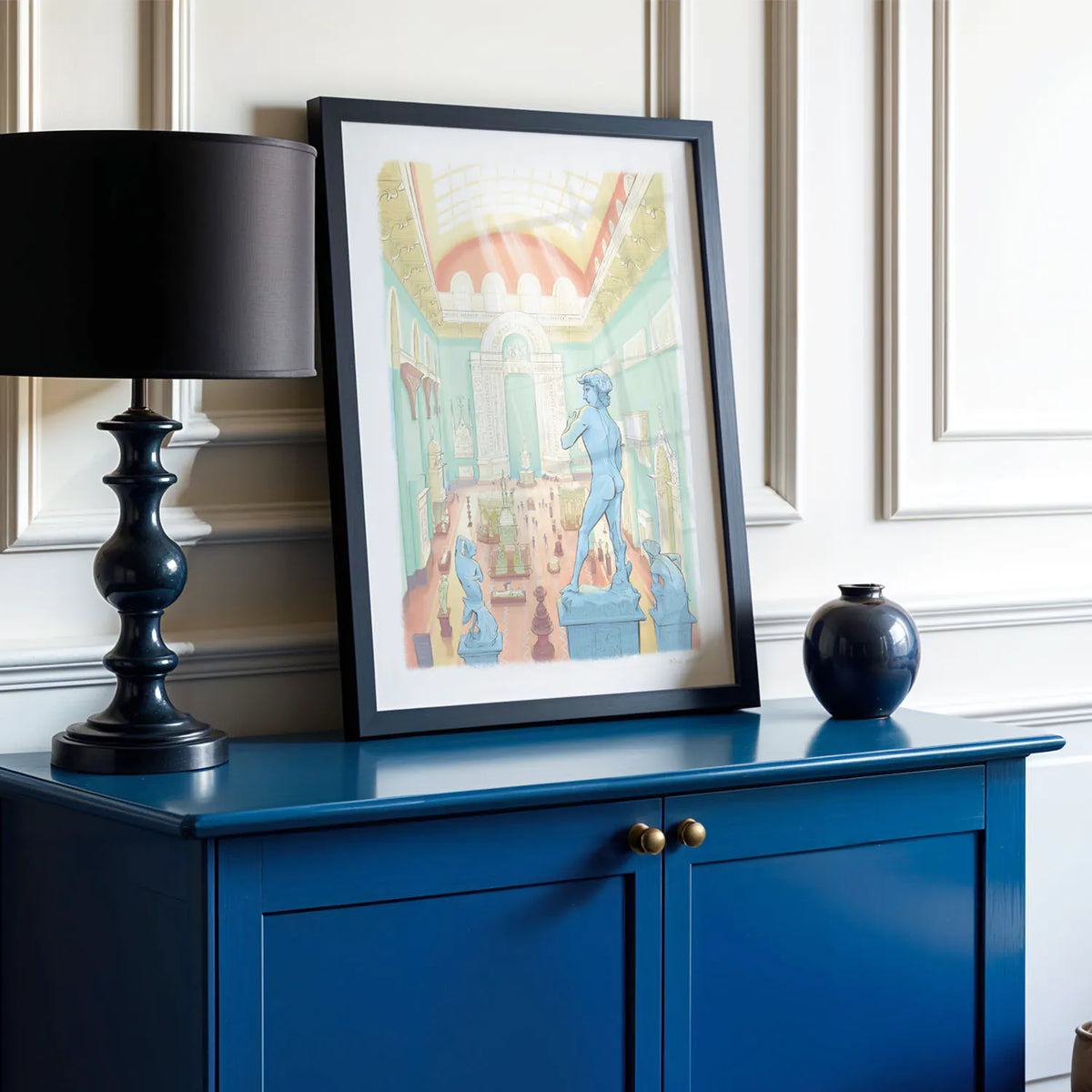 A framed art print featuring a colorful illustration of classical statues in an ornate interior, styled on a dark blue cabinet next to a black table lamp and a small dark vase against a white paneled wall.