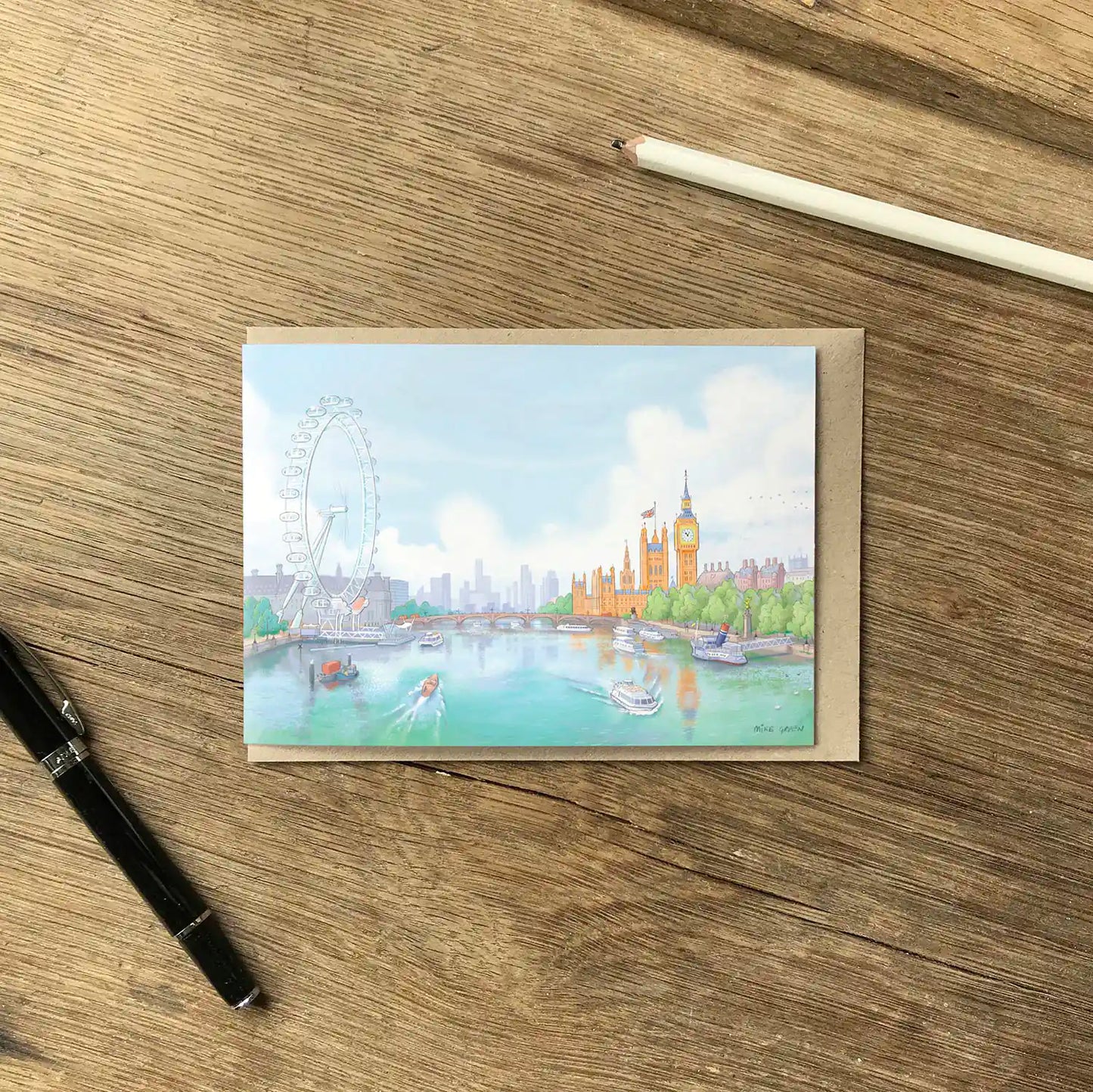 Greetings card on desk with pens of an illustration of the view south from London's Golden Greetings card of the view from Jubilee Bridge with Houses of Parliament and the London eye by Mike Green Illustration