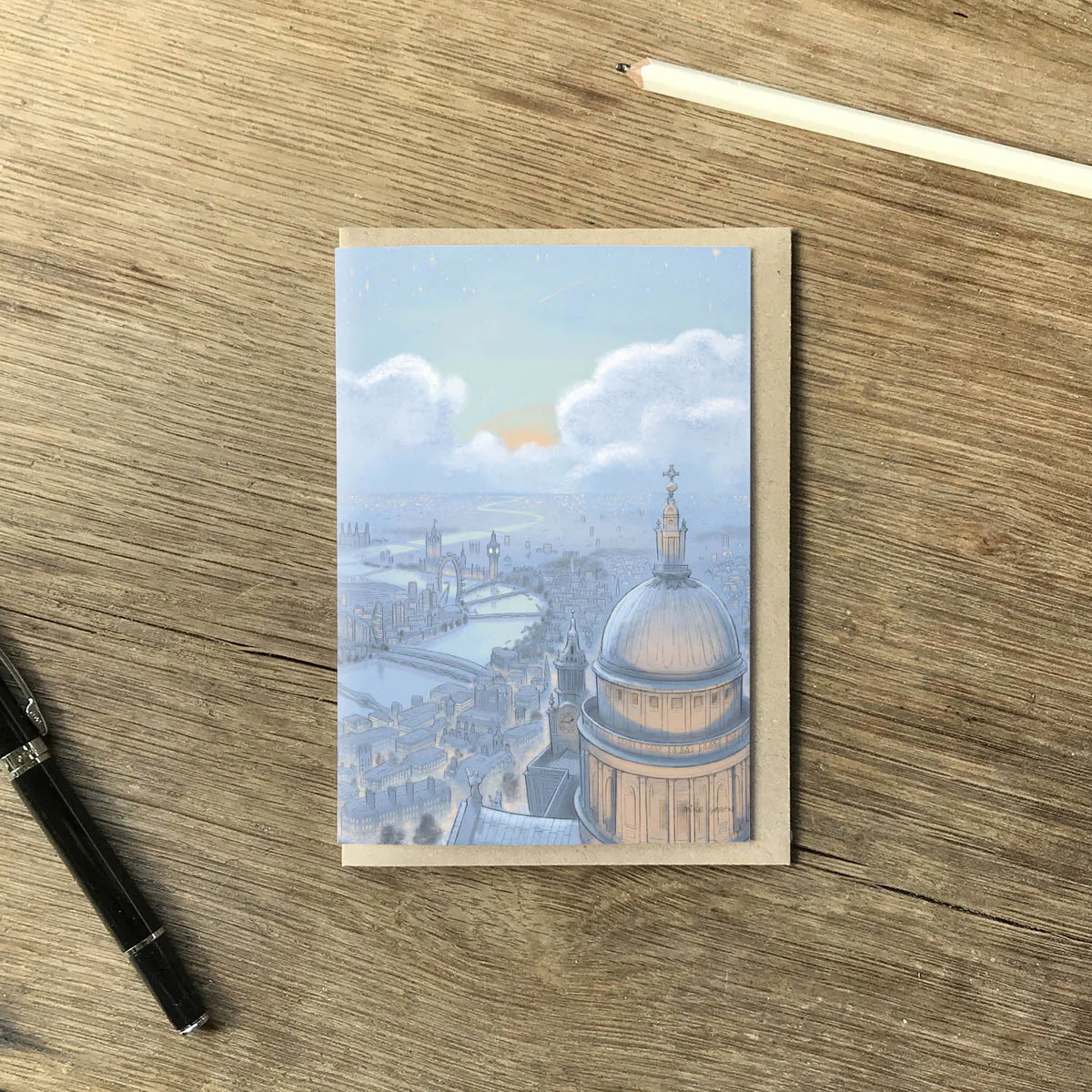 Greetings card with painting of London skyline at night with St Pauls Cathedral and Big Ben on a wooden table with a pencil and pen