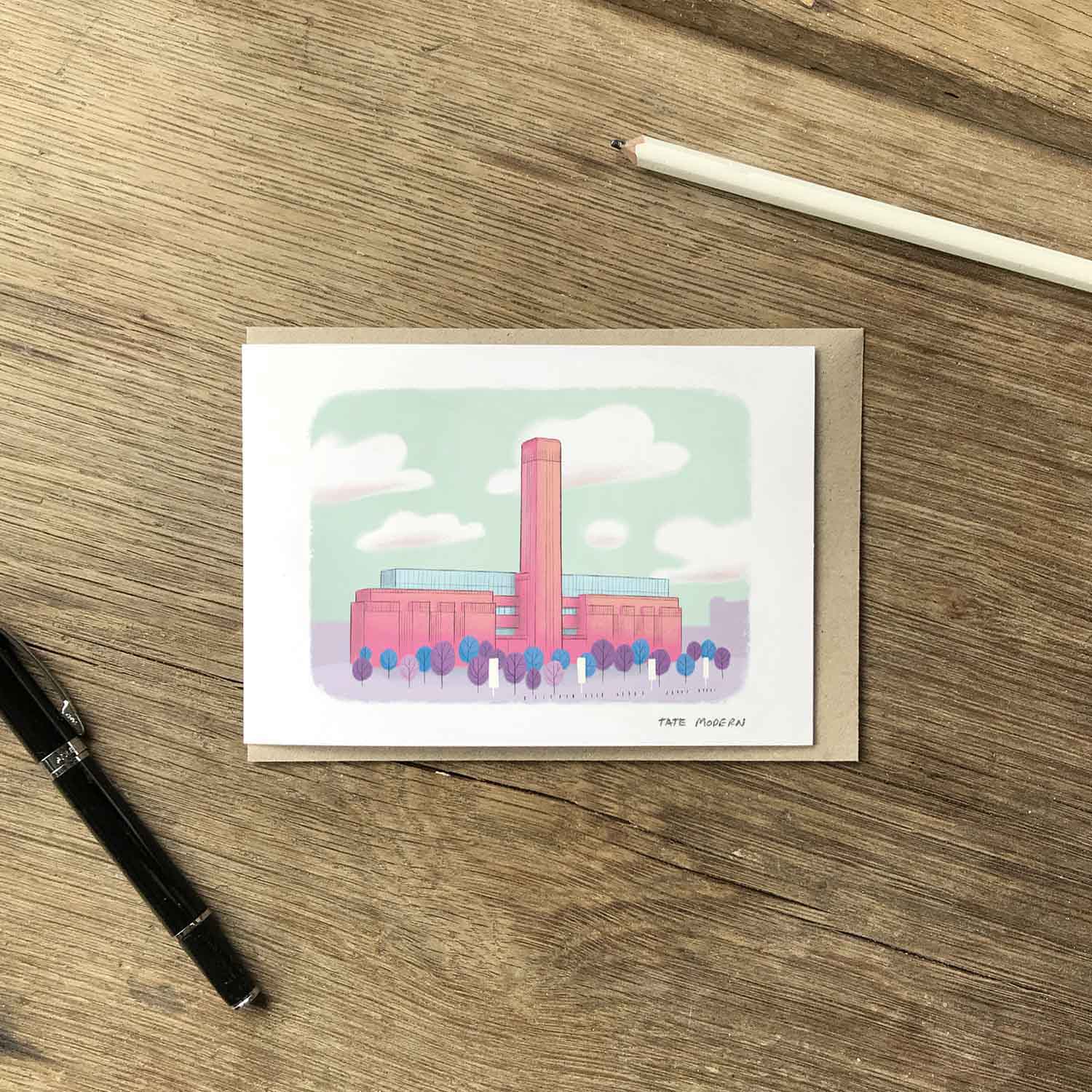 A greetings card with an illustration of London's Tate Modern gallery building, placed on a kraft envelope on a wooden tabletop with a pen and pencil nearby.