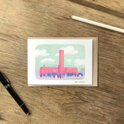 A greetings card with an illustration of London's Tate Modern gallery building, placed on a kraft envelope on a wooden tabletop with a pen and pencil nearby.