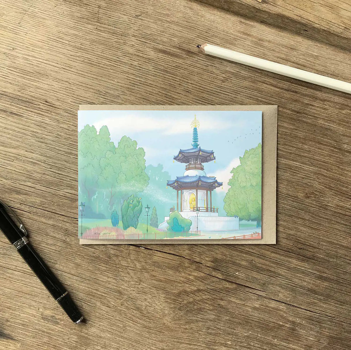 A greeting card showing a painted pagoda in London's Battersea Park, placed on a wooden tabletop with a pencil and pen nearby.