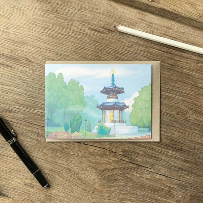 A greeting card showing a painted pagoda in London's Battersea Park, placed on a wooden tabletop with a pencil and pen nearby.