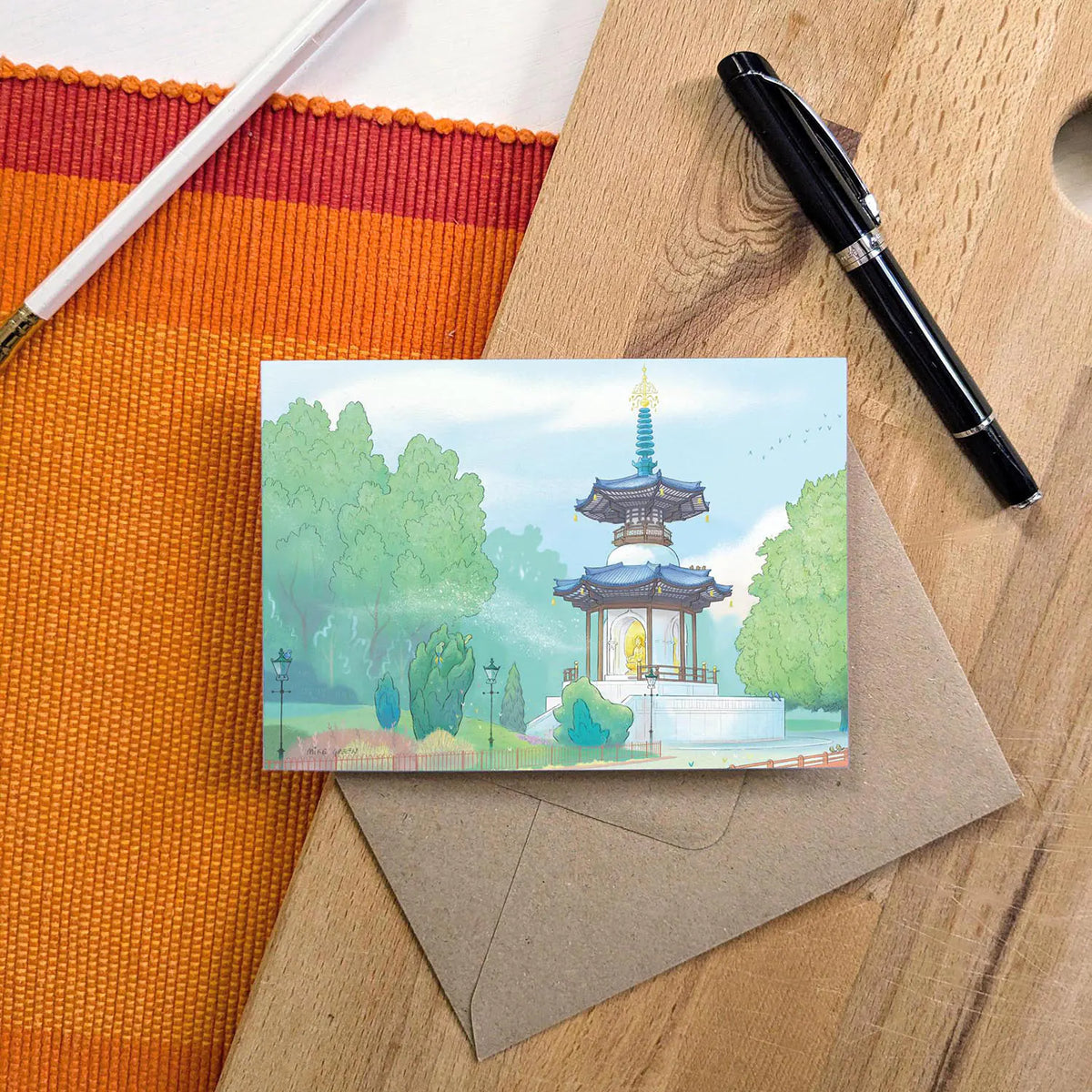London's Battersea Park Peace Pagoda illustrated on a greeting card with a pen and pencil with a kraft envelope on a wooden surface.