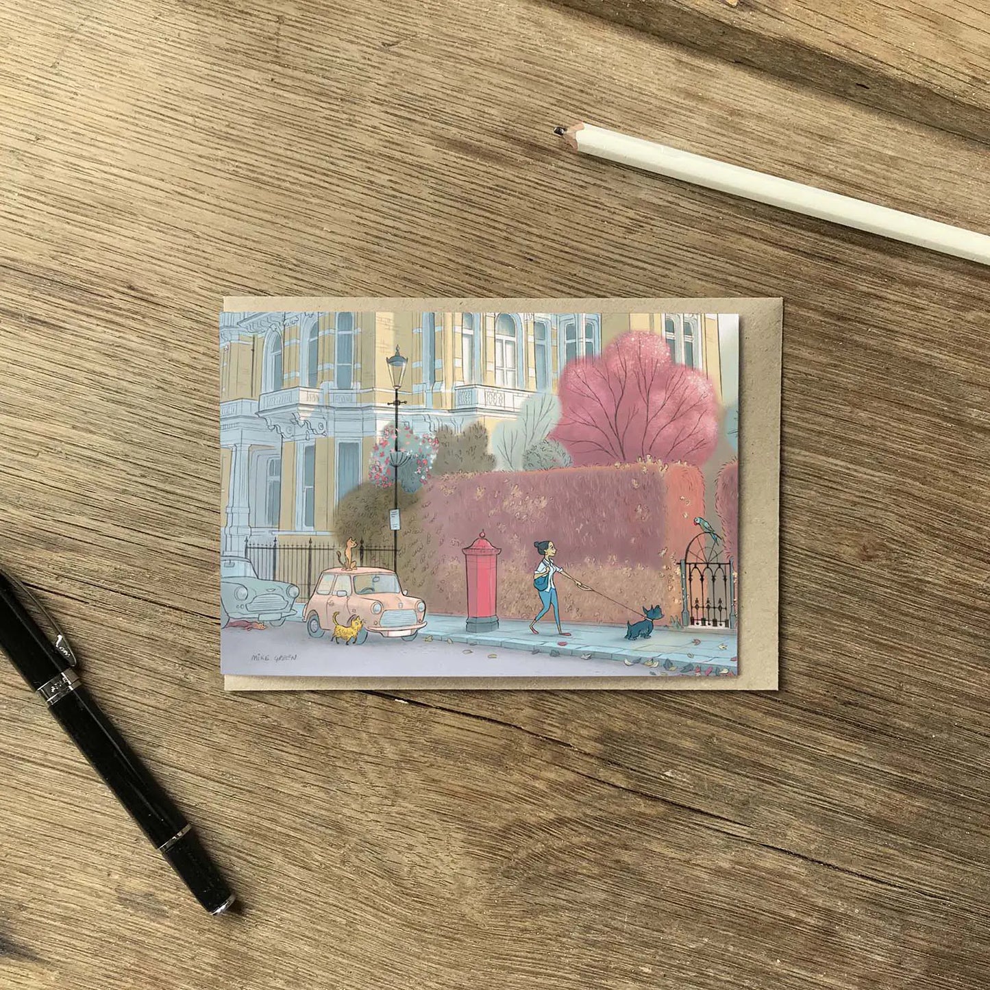 A lady and dog walking in london's Kensington street with a parakeet cars and cats on a street in London's Kensington in this illustrated greeting card shown with a pen and envelope on a wooden surface.
