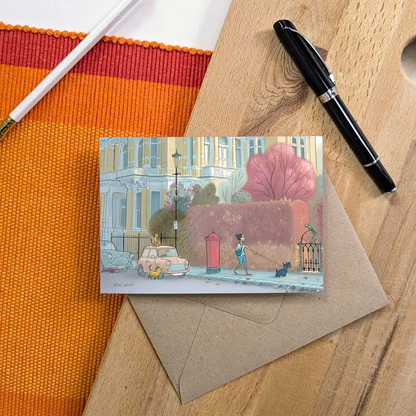A lady and dog walking in london's Kensington street with a parakeet cars and cats on a street in London's Kensington in this illustrated greeting card shown with a pen and envelope on a wooden surface.