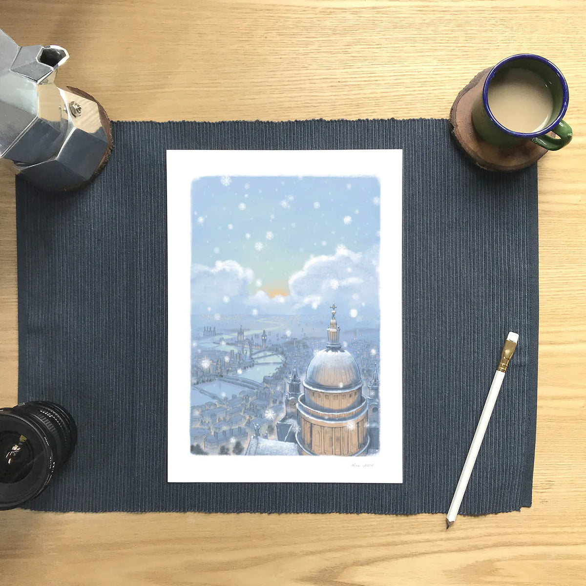 Wall art print of St Paul's Cathedral and London skyline in the evening with falling snow next to a pencil and cup of coffee