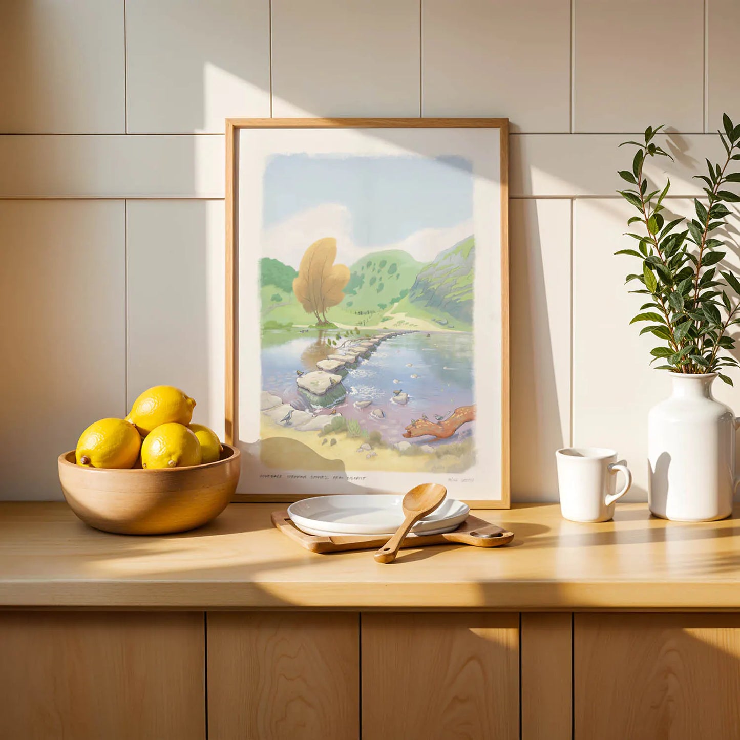 Framed wall art print of a painting of the stepping stones in Dovedale in the Peak District national park on a shelf with lemons