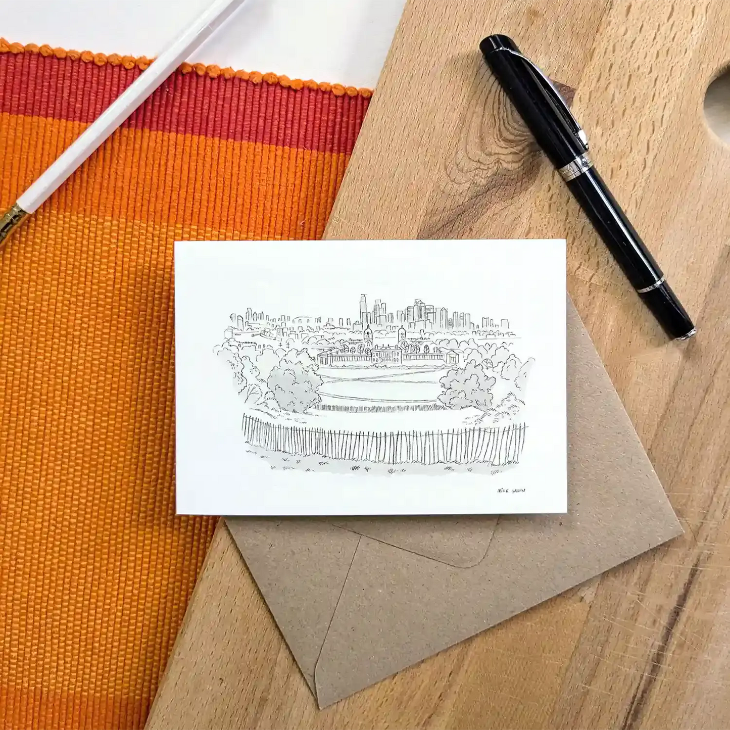 A white greeting card featuring a black ink sketch of Greenwich park and London skyline, placed on a brown kraft envelope on a wooden surface. A black pen and a white pencil are visible as props around it.