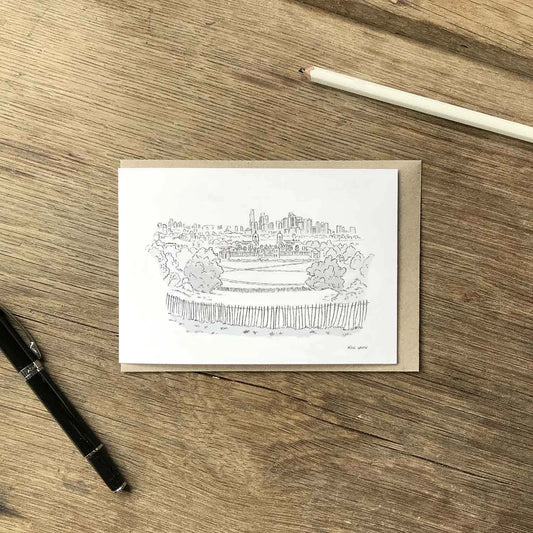 A white greeting card featuring a black ink sketch of Greenwich park and London skyline, placed on a brown kraft envelope on a wooden tabletop. A black pen and a white pencil are visible as props around it.