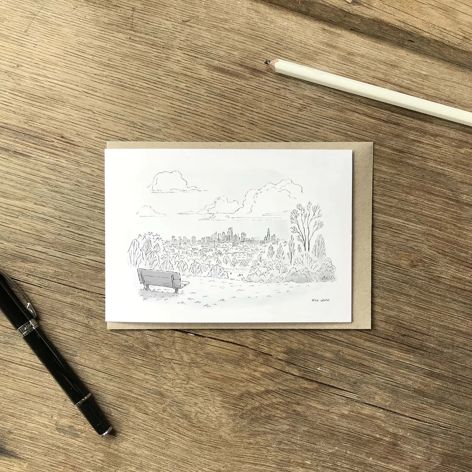 A greeting card featuring a black ink sketch of Hampstead Heath in north London, placed on top of a brown kraft envelope on a wooden surface with a white pencil, and a black pen as props.