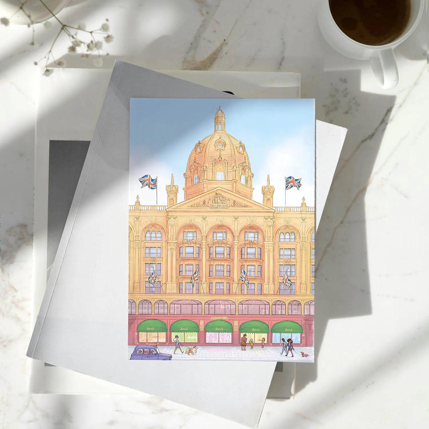 A printed card of an illustration of Harrods store in London's Knightsbridge resting on a pile of books on a white marble surface with a coffee cup nearby.