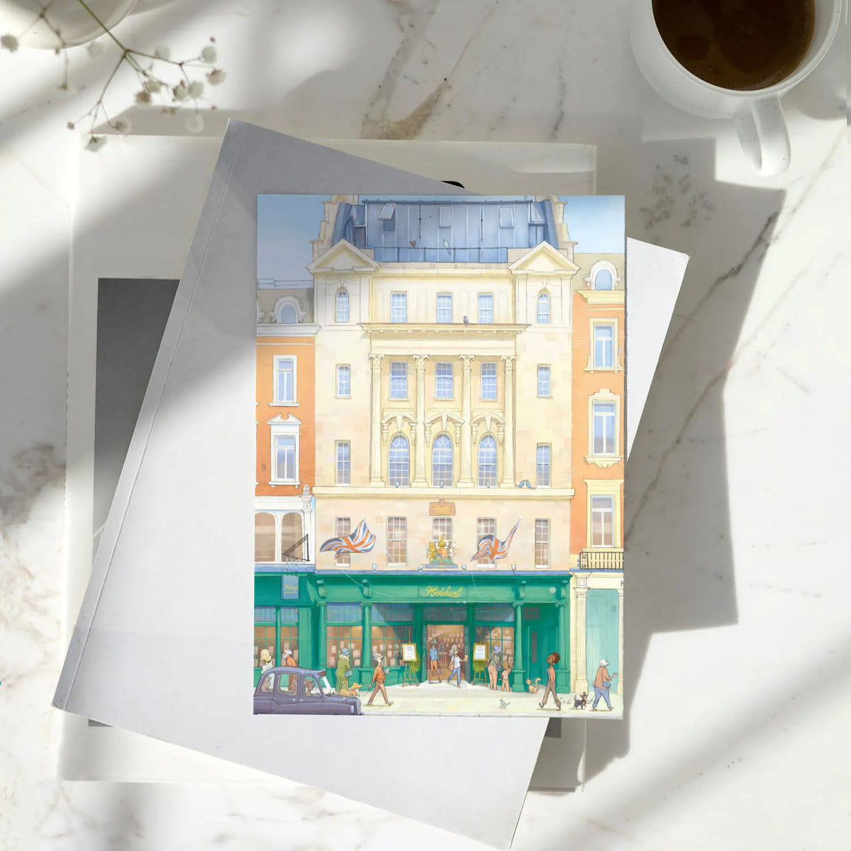 A greetings card featuring a watercolor-style illustration of Hatchards Piccadilly bookshop in London with a kraft envelope behind it on a pile of books on a marble surface