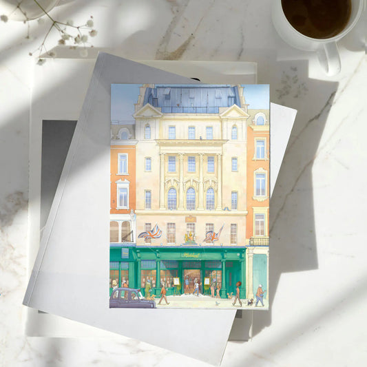 A greetings card featuring a watercolor-style illustration of Hatchards Piccadilly bookshop in London with a kraft envelope behind it on a pile of books on a marble surface