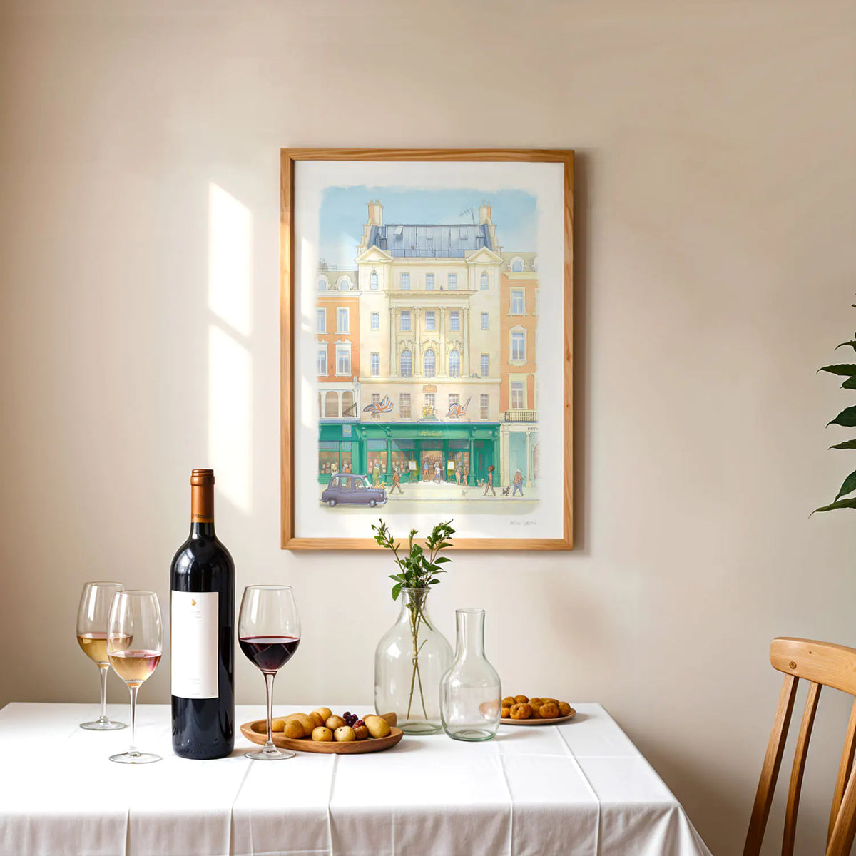 A framed watercolor wall art print of Hatchards book shop in London, hung on a beige wall above a dining table set with a bottle of red wine, glasses, and small plates of food.