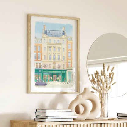 A framed watercolor wall art print of Hatchards book shop in London hangs on a beige wall above a wooden console table styled with books, a ceramic vase, and a mirror.