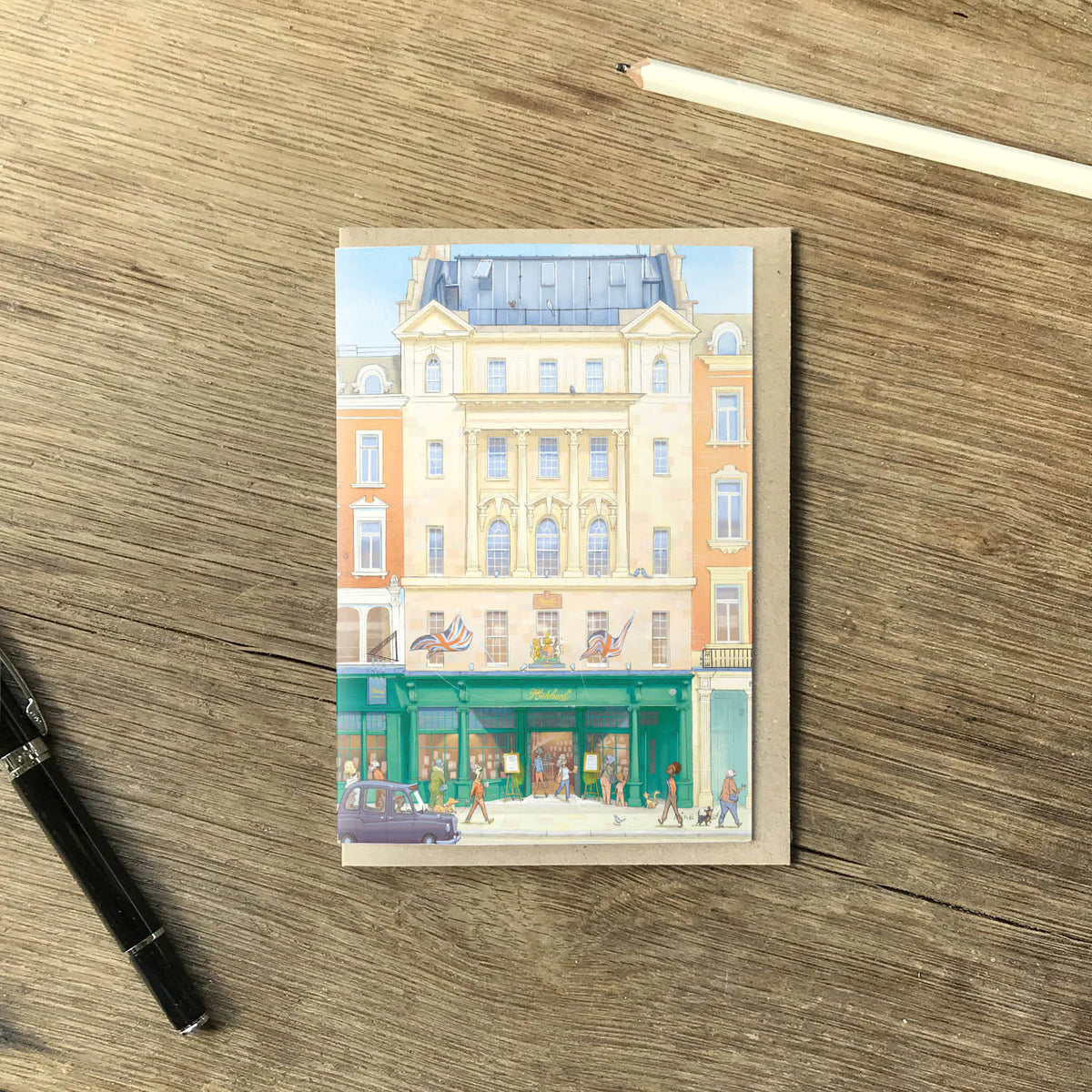 A greetings card featuring a watercolor-style illustration of Hatchards Piccadilly bookshop in London with a kraft envelope behind it on a wooden surface with a pencil and pen.
