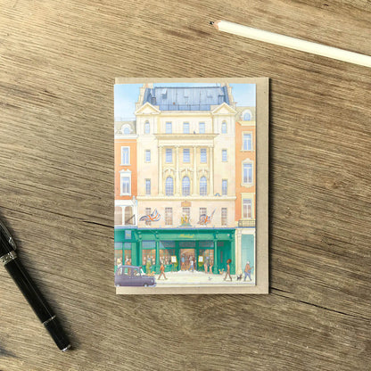 A greetings card featuring a watercolor-style illustration of Hatchards Piccadilly bookshop in London with a kraft envelope behind it on a wooden surface with a pencil and pen.
