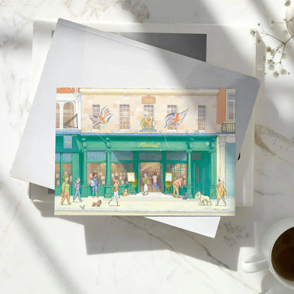 A greetings card featuring a watercolor-style illustration of Hatchards Piccadilly bookshop in London with a kraft envelope behind it on a pile of books on a marble surface