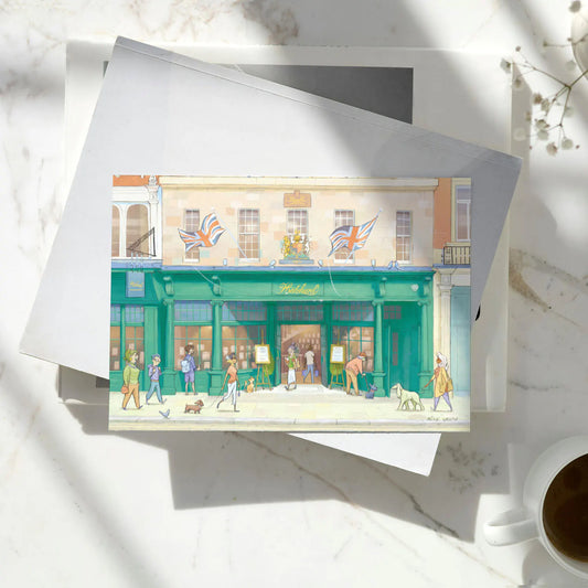 A greetings card featuring a watercolor-style illustration of Hatchards Piccadilly bookshop in London with a kraft envelope behind it on a pile of books on a marble surface