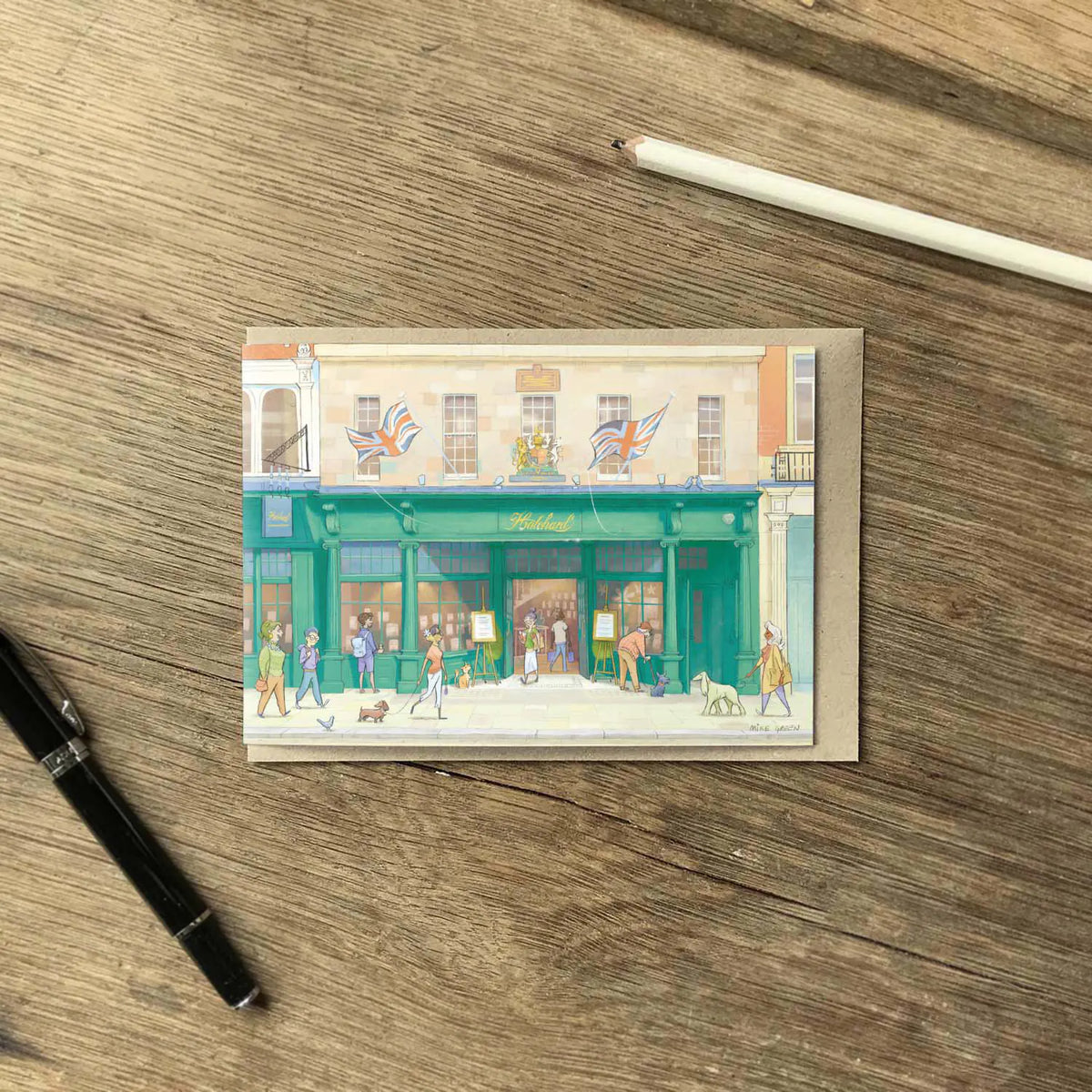 A greetings card featuring a watercolor-style illustration of Hatchards Piccadilly bookshop in London with a kraft envelope behind it on a wooden surface with a pencil and pen.
