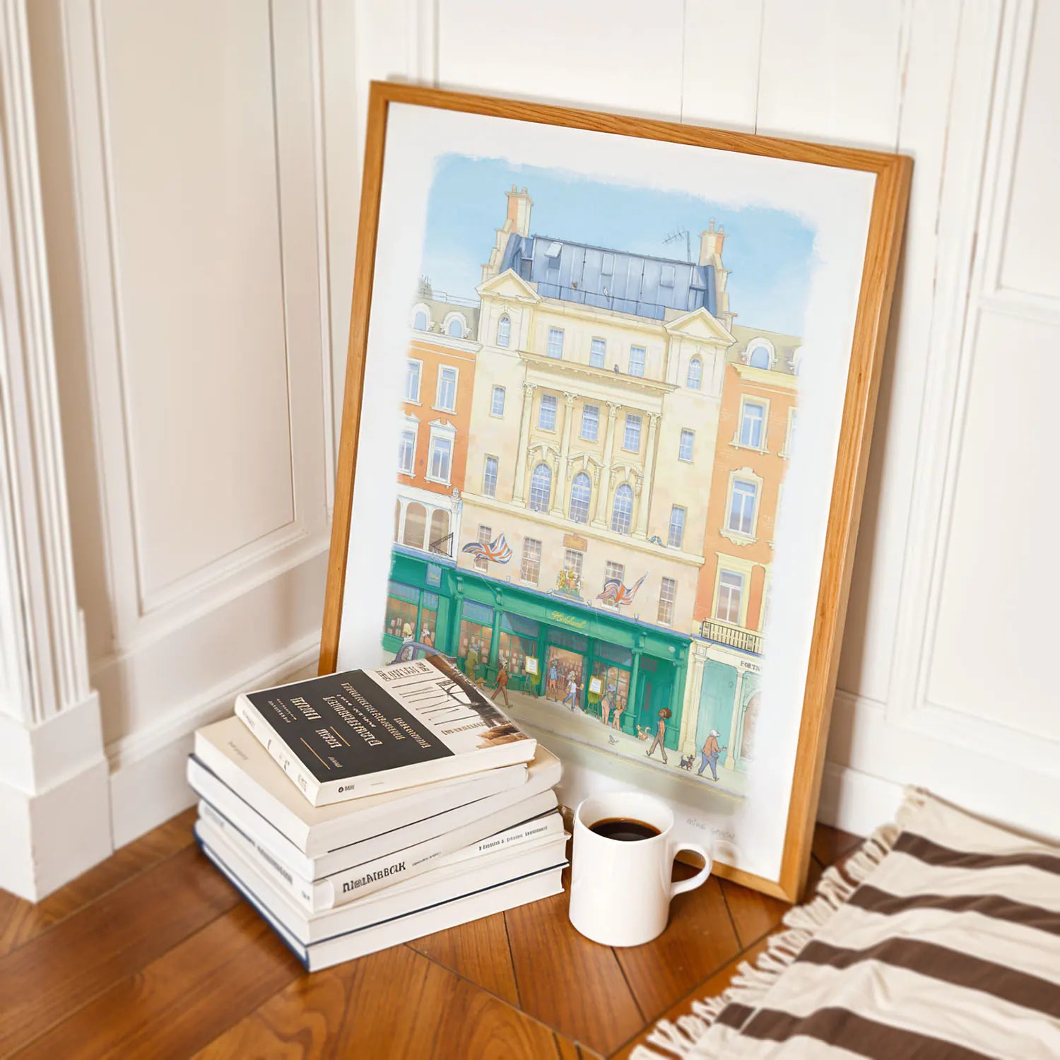 A framed watercolor wall art print of Hatchards book shop in London leans against a white paneled wall on a wooden floor, styled next to a stack of books and a white mug.
