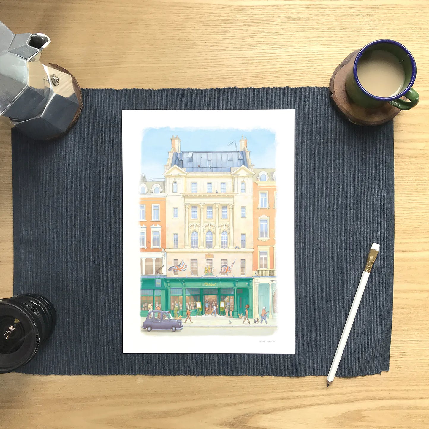 A wall art print of Hatchards bookshop in Piccadilly London in an illustration water colour style, displayed on a dark navy ribbed placemat over a light wood desk, surrounded by coffee and study props.