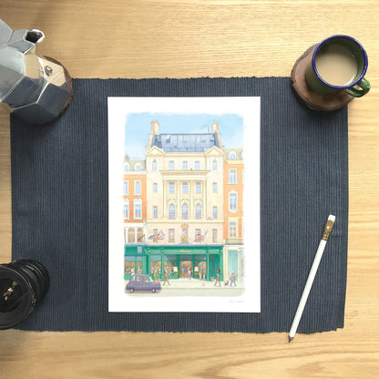 A wall art print of Hatchards bookshop in Piccadilly London in an illustration water colour style, displayed on a dark navy ribbed placemat over a light wood desk, surrounded by coffee and study props.