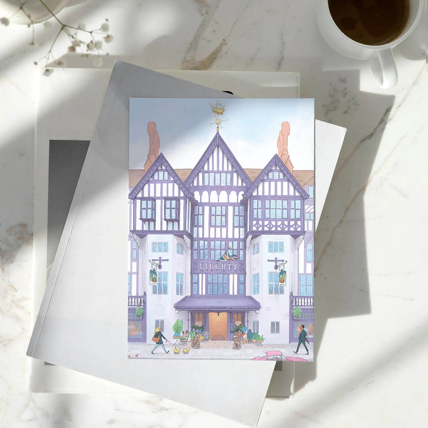 London's Liberty Store illustrated on a greeting card resting on a pile of books on a marble surface with a coffee cup.