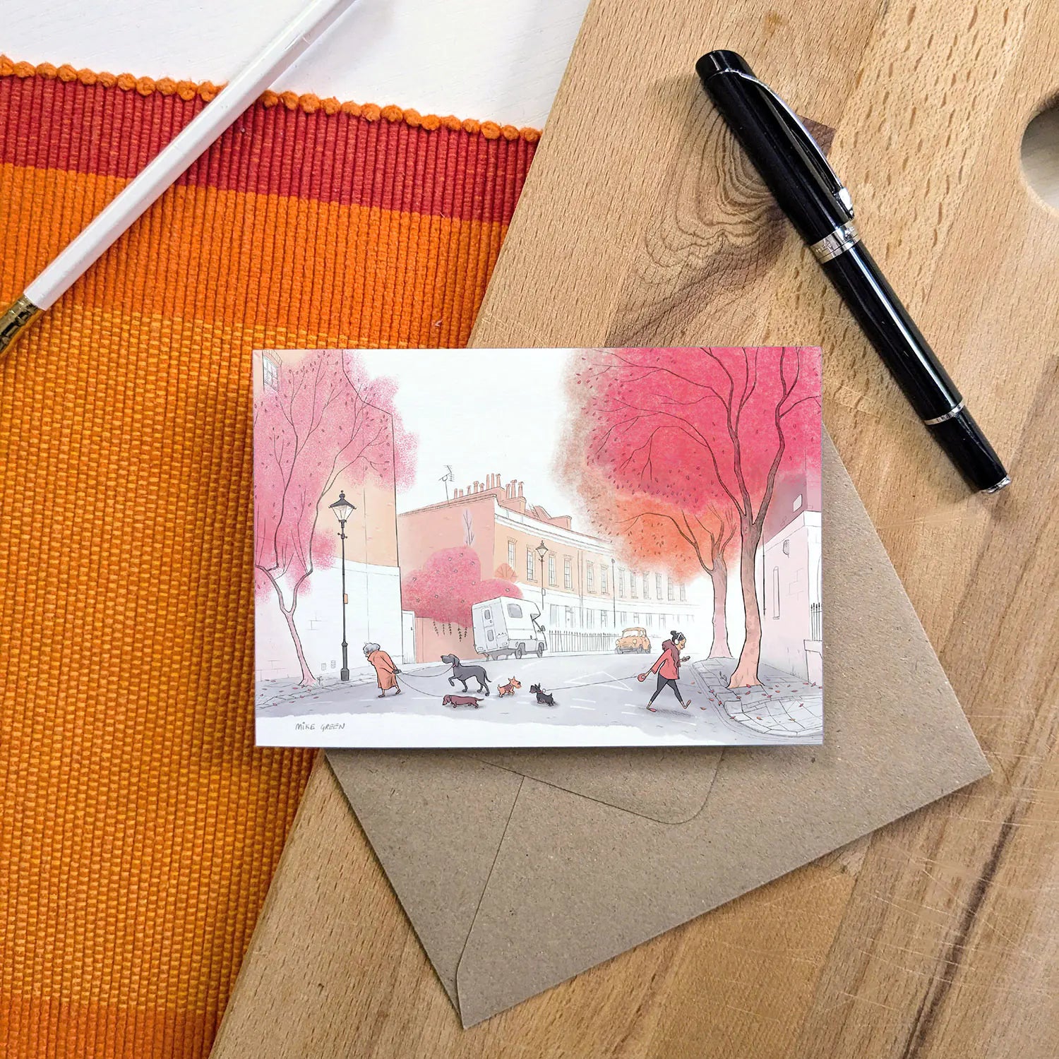 A greetings card with an illustration of dogs in the autumn on a street in london with a pen and envelope on a wooden surface.