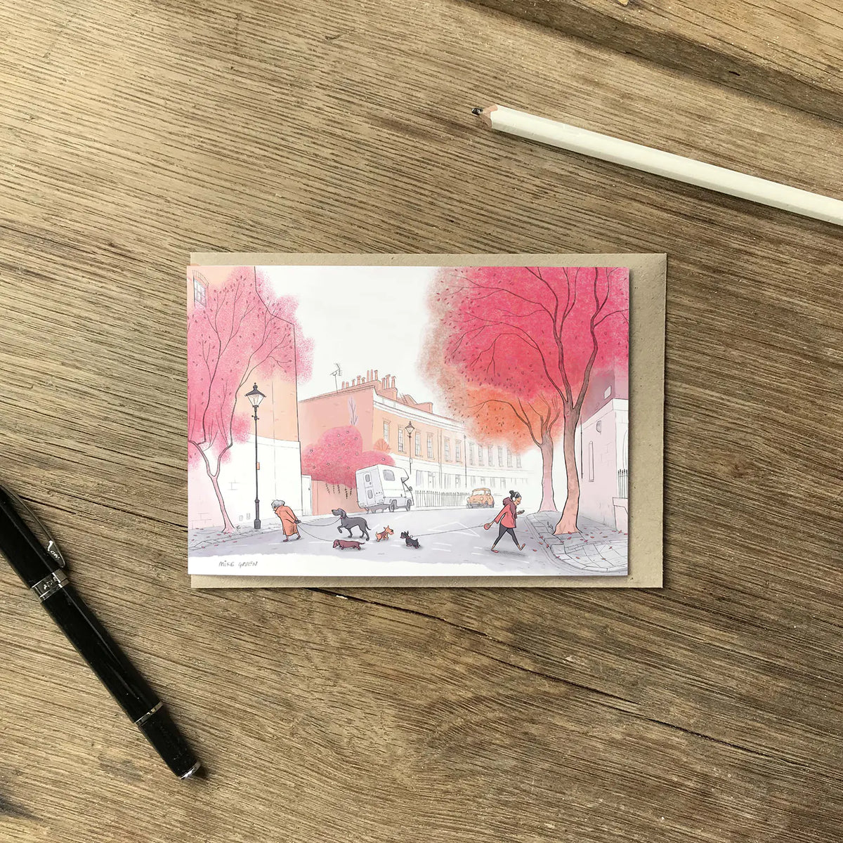 A greetings card with an illustration of dogs in the autumn on a street in london with a pen and envelope on a wooden surface.