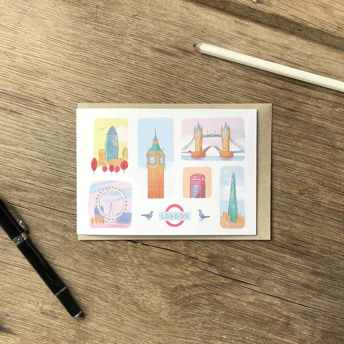 A rectangular greeting card with a kraft envelope, laid flat on a wooden tabletop. The card features six colorful illustrated London landmarks, including Big Ben, Tower Bridge, the London Eye, and the London Underground roundel.