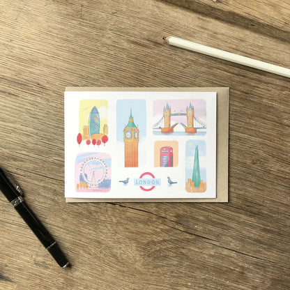 A rectangular greeting card with a kraft envelope, laid flat on a wooden tabletop. The card features six colorful illustrated London landmarks, including Big Ben, Tower Bridge, the London Eye, and the London Underground roundel.