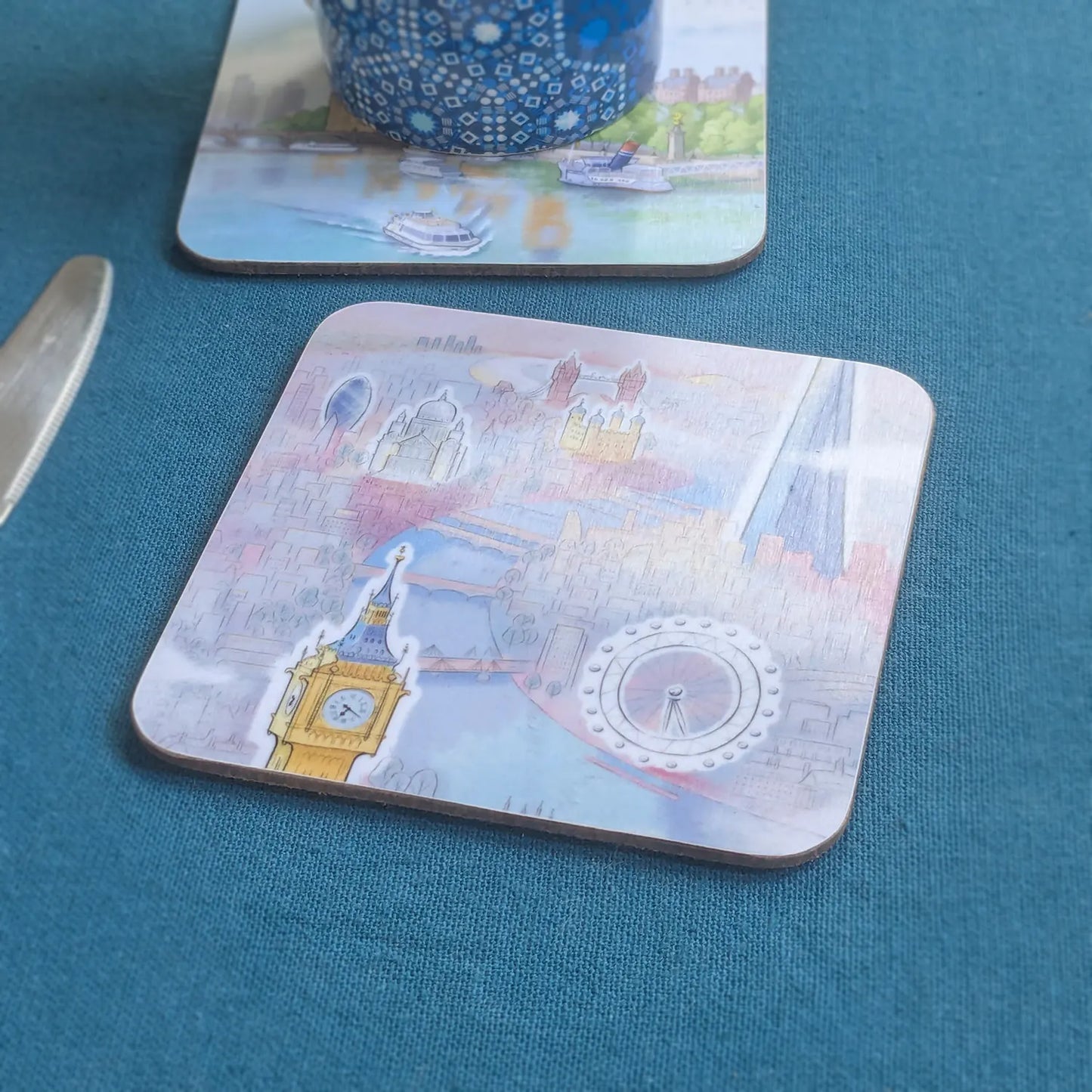 A close-up of a square coaster with rounded corners featuring a London-themed collage design, resting on a textured teal tablecloth next to a knife. Another coaster and a blue patterned mug are partially visible in the background.