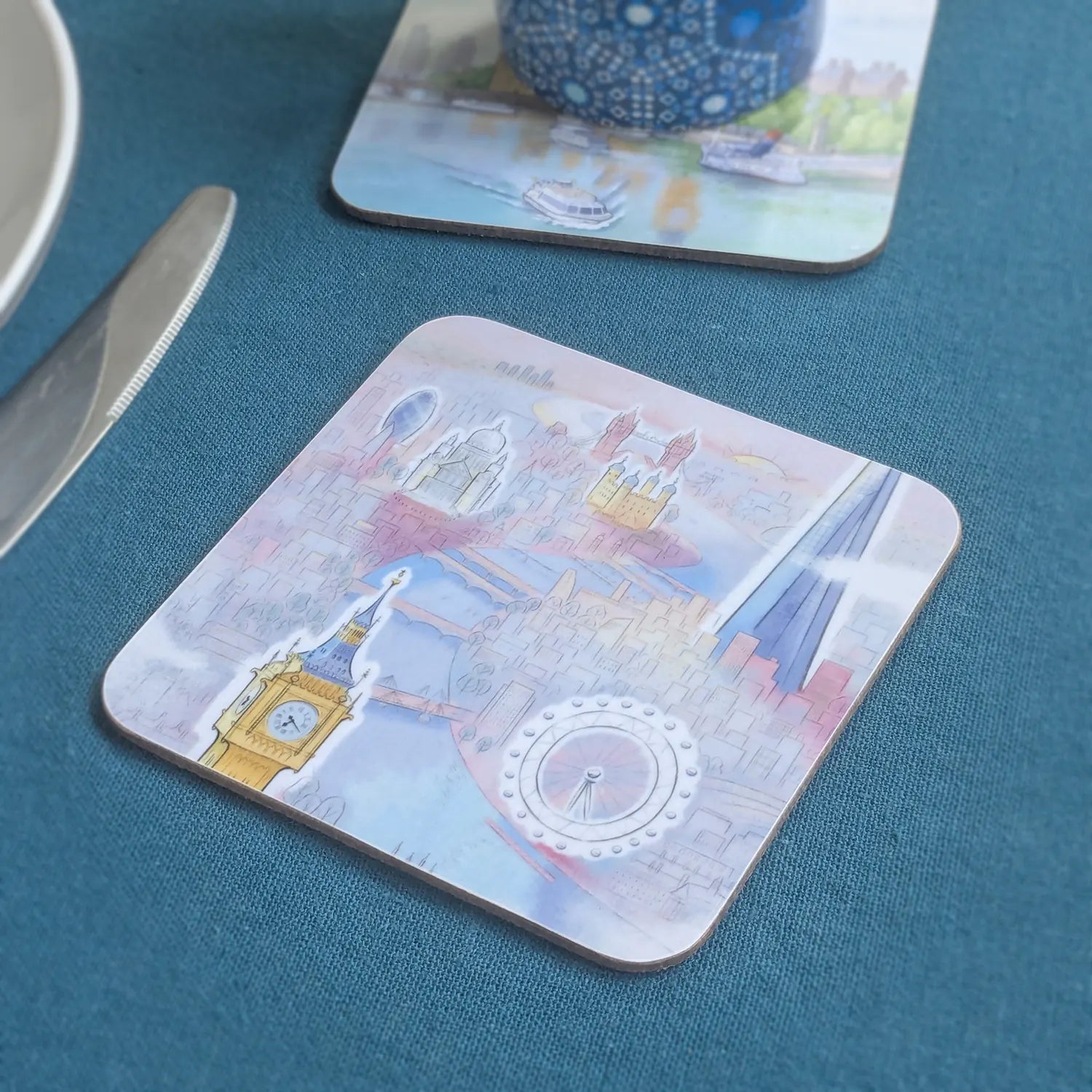 Square cork drinks coaster with rounded corners of London skyline in pastel shades on a table cloth with coaster in background