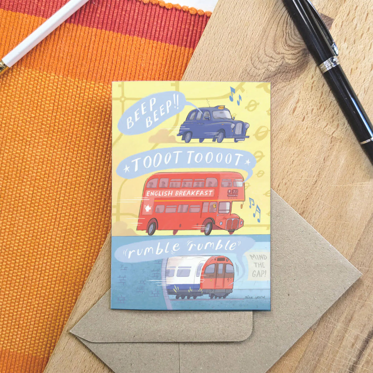 A greetings card with London transport, a red bus, a taxi and a tube train all making sounds with an envelope and pen on a wooden board