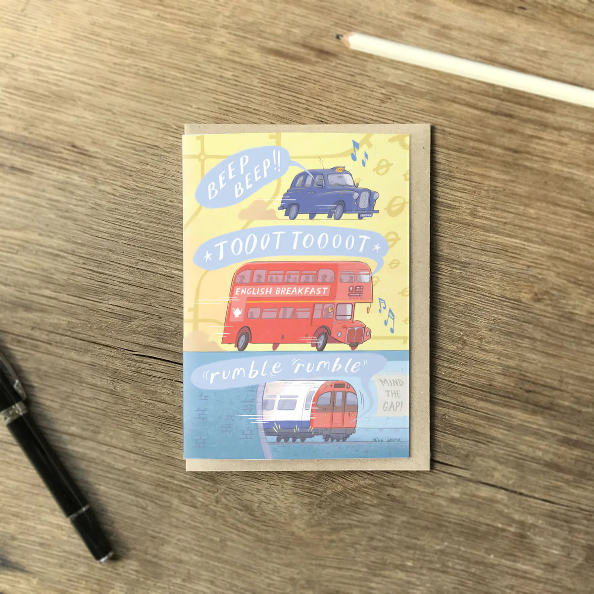 A greetings card with London transport, a red bus, a taxi and a tube train all making sounds with an envelope and pencil on a wooden surface