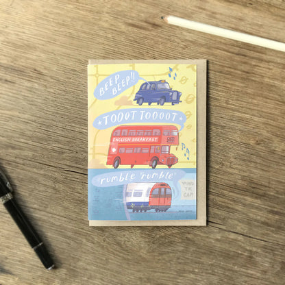 A greetings card with London transport, a red bus, a taxi and a tube train all making sounds with an envelope and pencil on a wooden surface