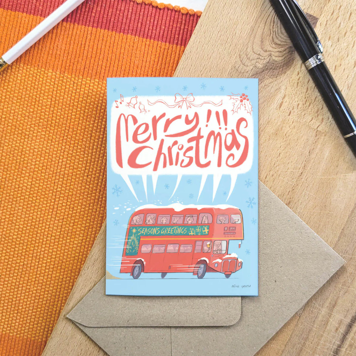 A Christmas greeting card featuring a red double-decker bus and the words 'Merry Christmas' in red script on a light blue background, placed on top of a kraft envelope over a wood surface with an orange woven mat and pens nearby.