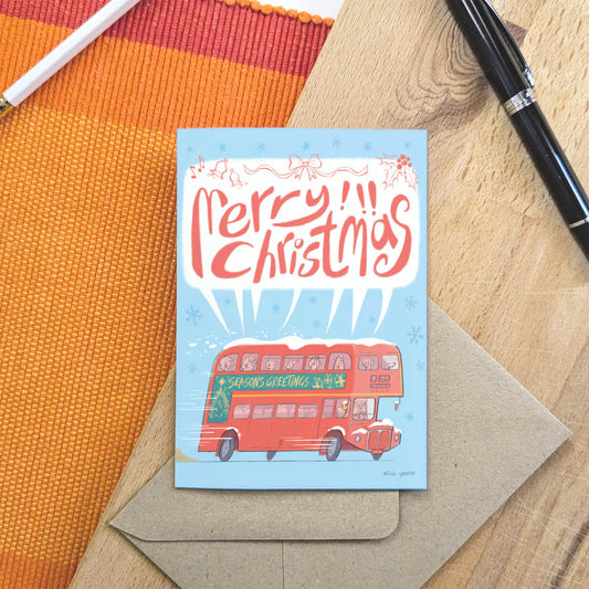 A Christmas greeting card featuring a red double-decker bus and the words 'Merry Christmas' in red script on a light blue background, placed on top of a kraft envelope over a wood surface with an orange woven mat and pens nearby.
