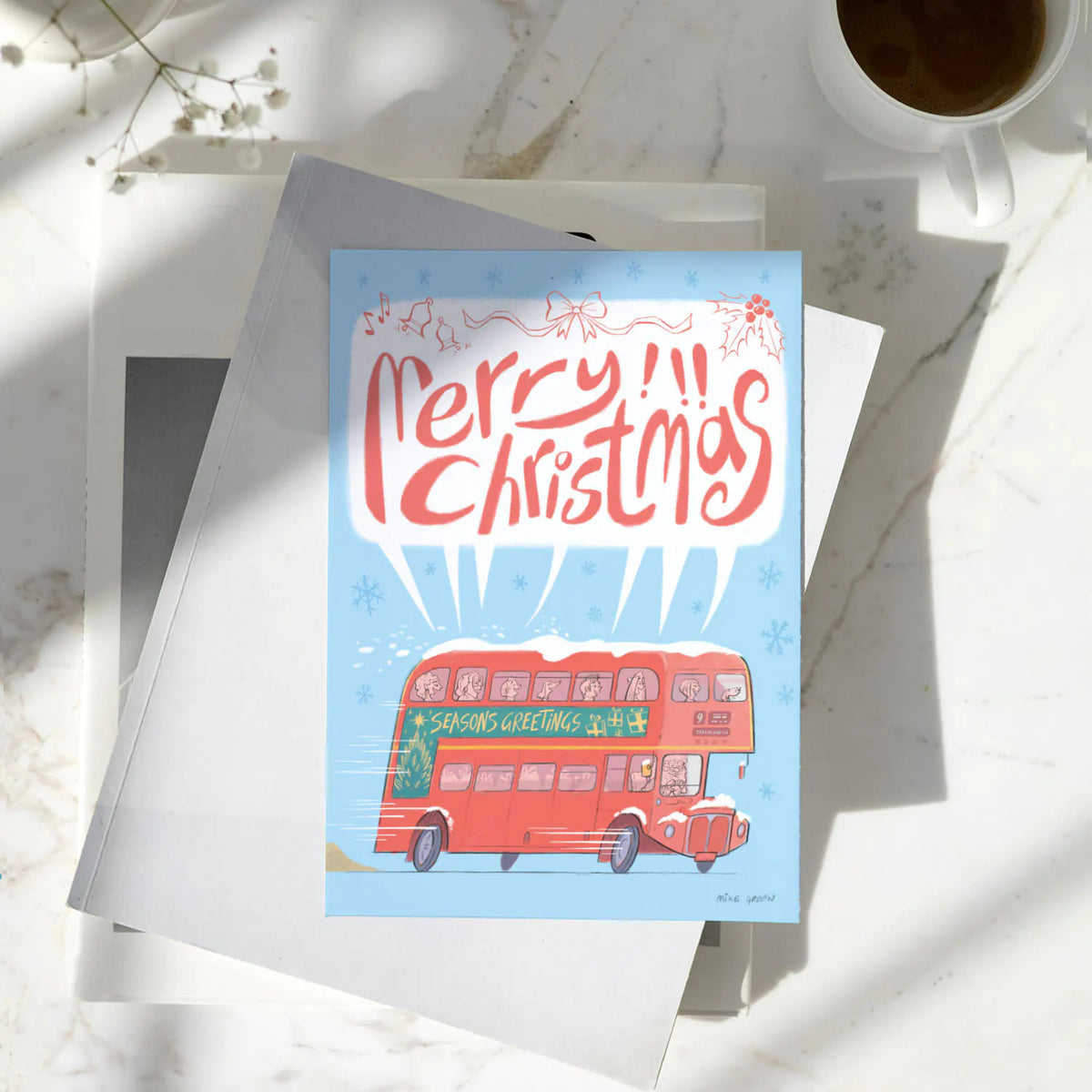 A Christmas greeting card featuring a red double-decker bus and the text 'Merry Christmas' on a light blue background, styled on top of white envelopes and a white marble surface with a coffee cup nearby.