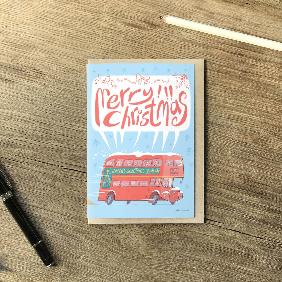 A rectangular Christmas greeting card lying on a wooden tabletop, with a red double-decker bus and the words “Merry Christmas” printed on the front. A black pen is at lower left, a pencil is at upper right, and no person or animal is present.