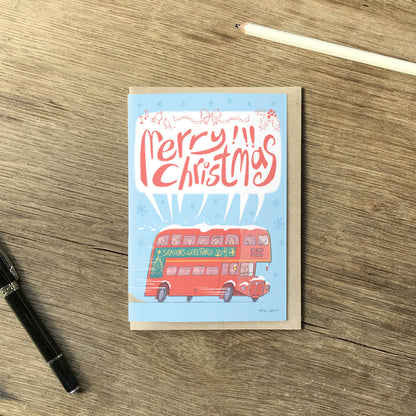 A rectangular Christmas greeting card lying on a wooden tabletop, with a red double-decker bus and the words “Merry Christmas” printed on the front. A black pen is at lower left, a pencil is at upper right, and no person or animal is present.