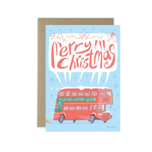 A Christmas greeting card with a kraft envelope behind it. The card features a light blue background, red hand-lettered text reading 'Merry Christmas,' and an illustration of a red double-decker bus with snow on top and 'Season's Greetings' written on the side.
