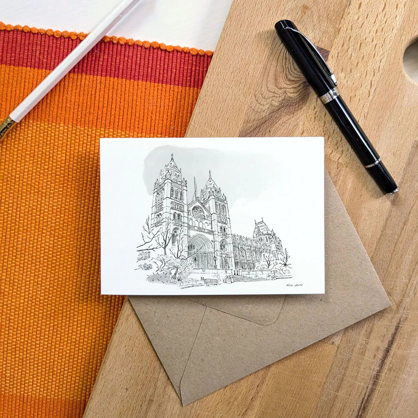 A white greeting card featuring a sketch of London's Natural History Museum in Kensington, displayed on a brown kraft envelope over a wooden desk with an orange striped mat, a white pencil, and a black pen.